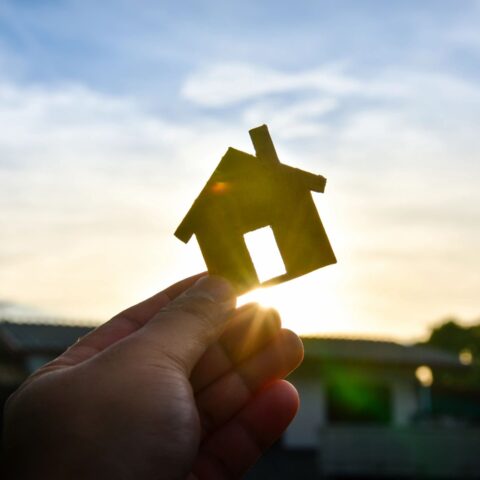 Image of hand holding a small wooden house cutout in front of a sunset